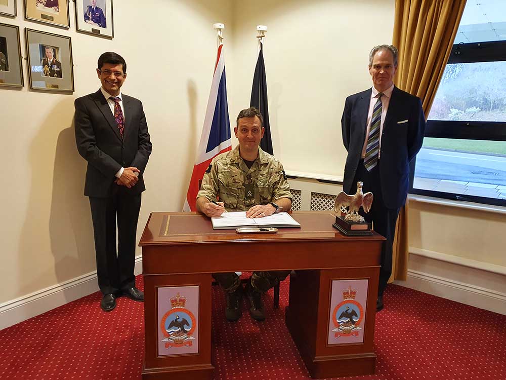 Army officer sat at a desk signing KIASS contract bookended by two individuals in suit and tie. 
