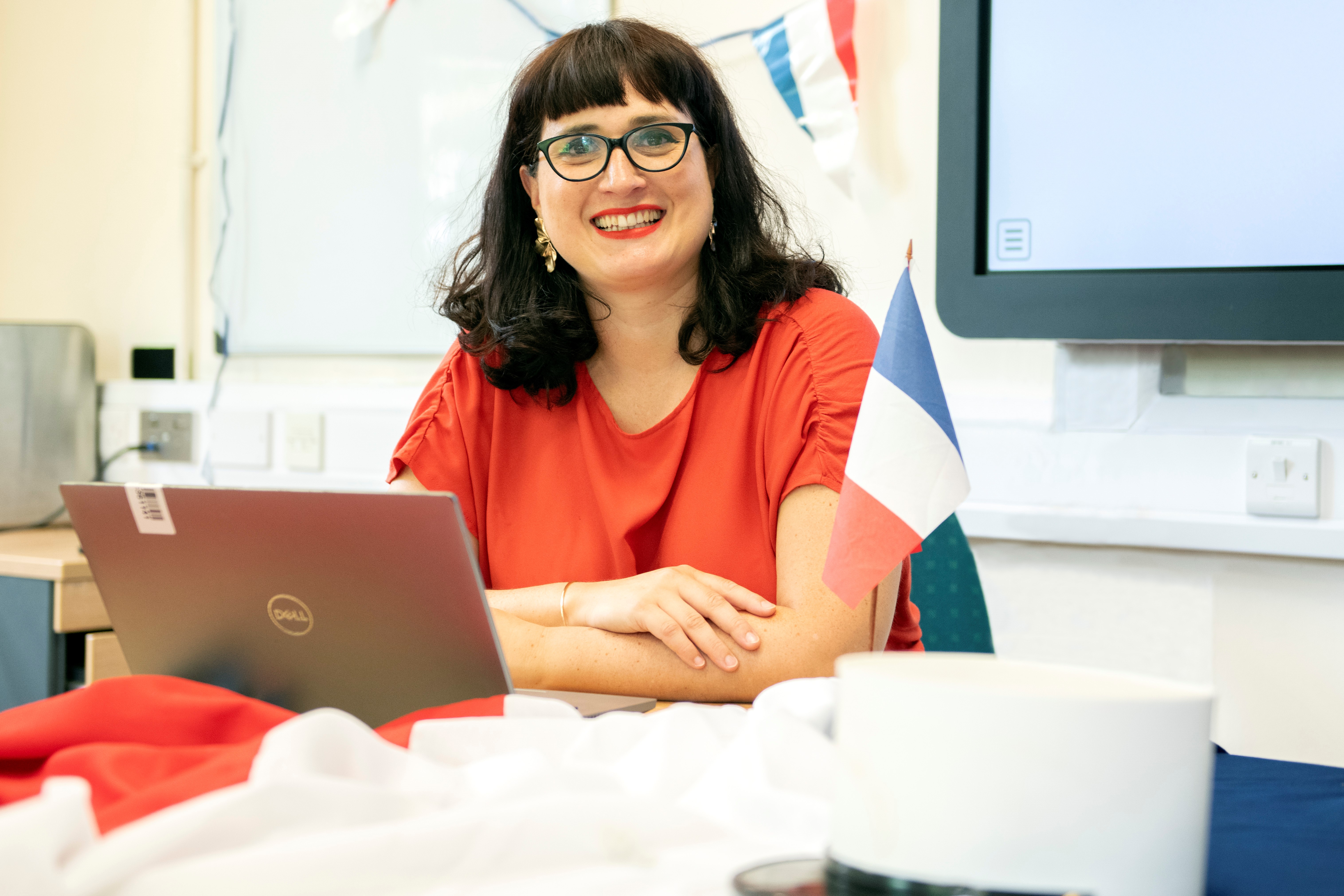 A woman in a red top and glasses sat at a desk with red, white and blue bunting in the background and a French flag on the desk.
