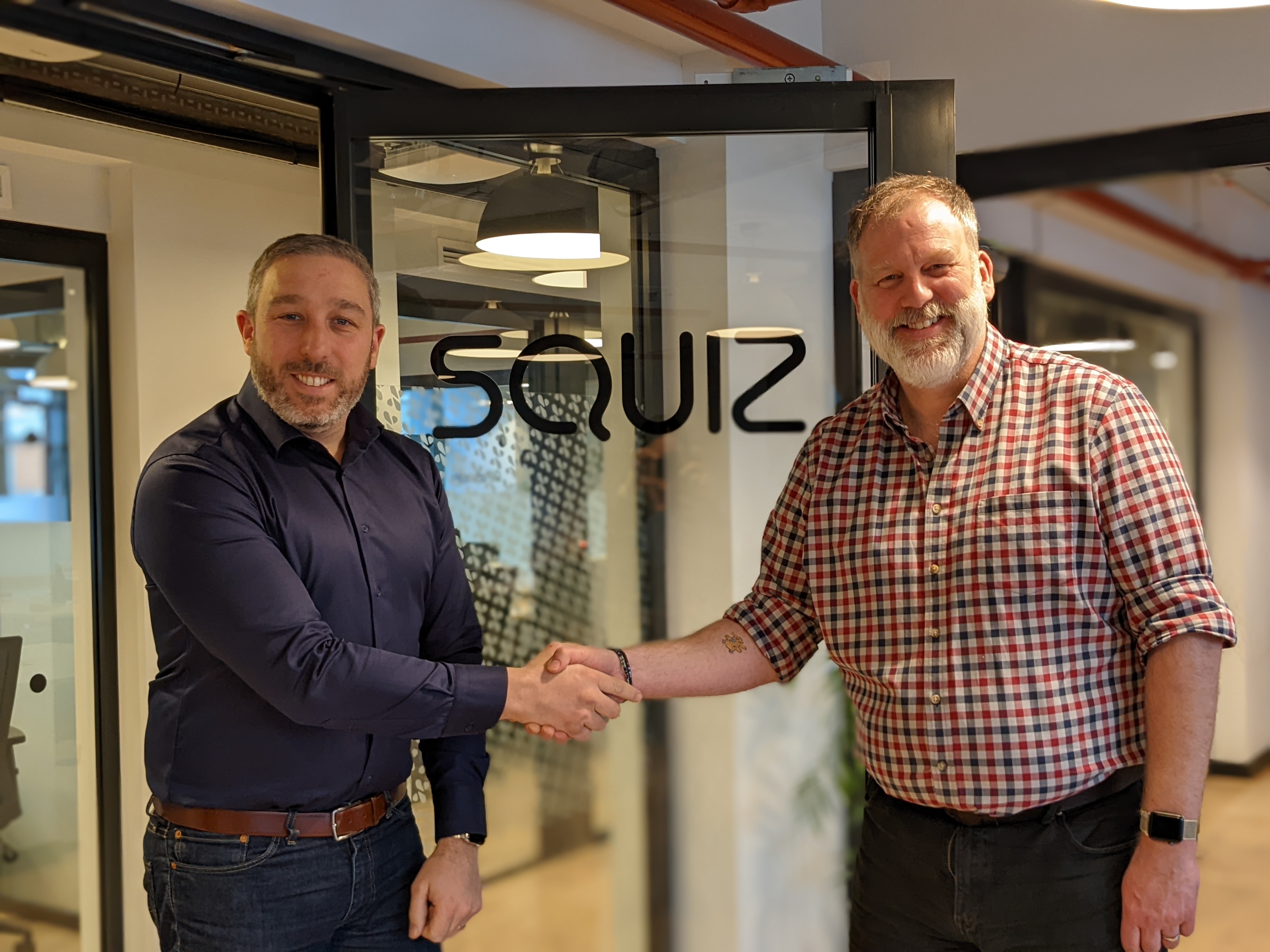 Two men in civilian dress shaking hands in front of a glass door with the company name 'Squiz'.