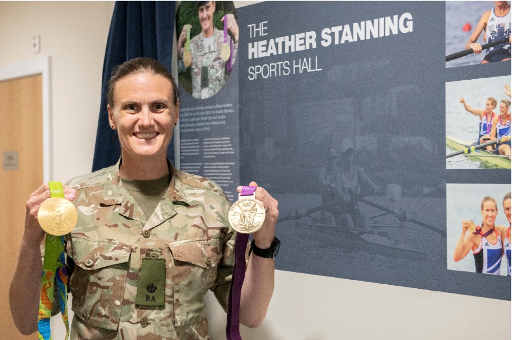 Heather Stanning holding her Olympic medals in front of her wall plaque inside the sports hall.