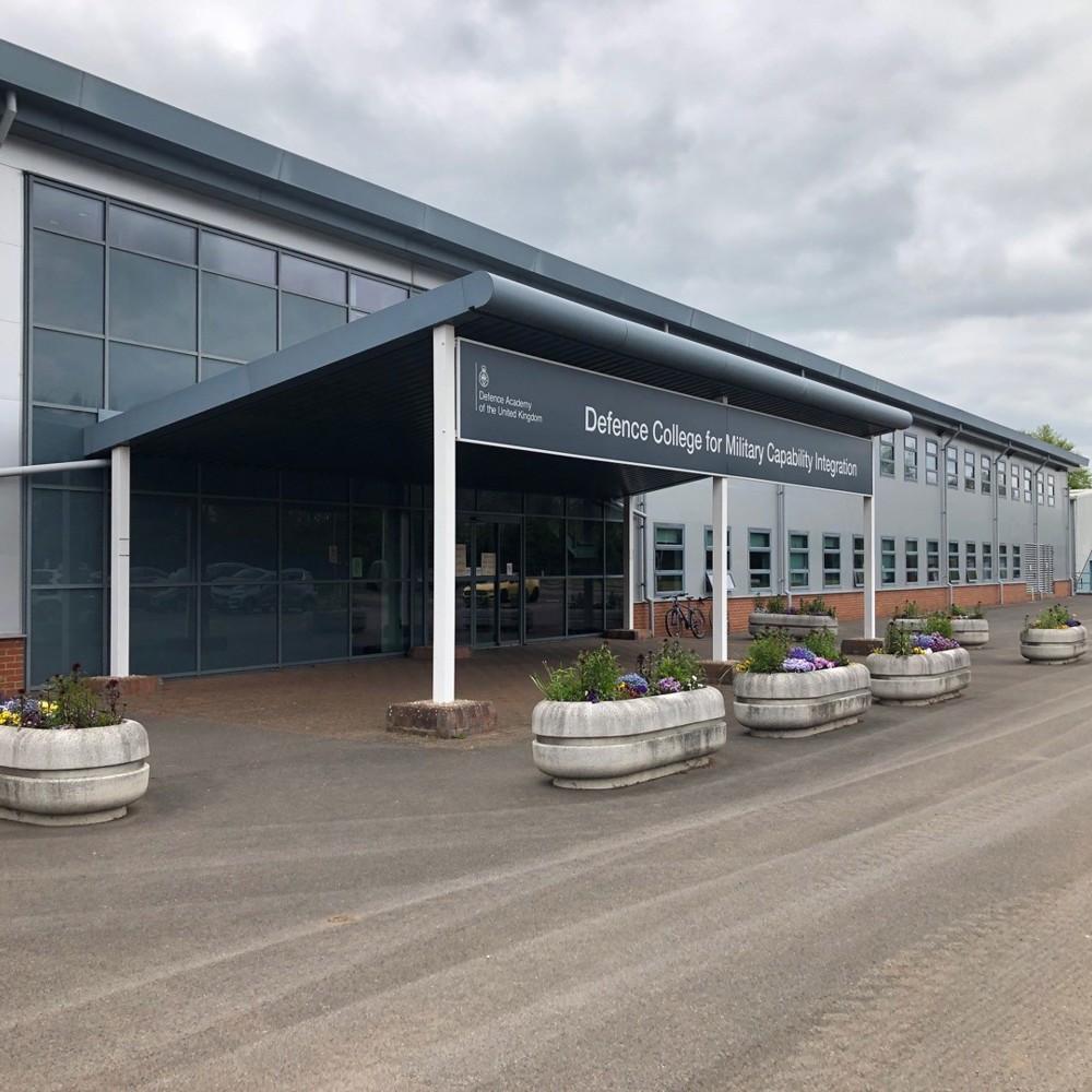 Entrance to a large building showing Defence College for Military Capability Integration on the signage with large concrete planters with colourful flowers at the doors. 