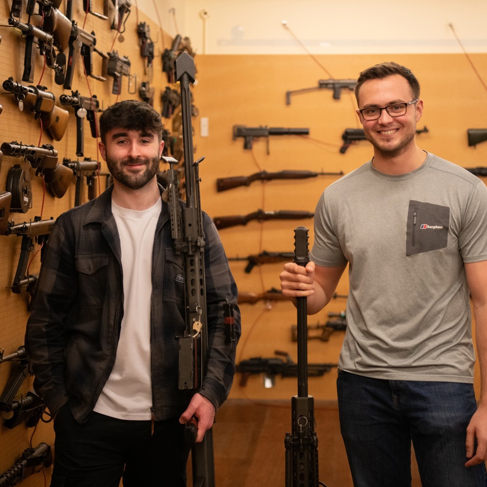 Two men stand in a room filled with various rifles and firearms displayed on wooden walls, each holding a large gun.