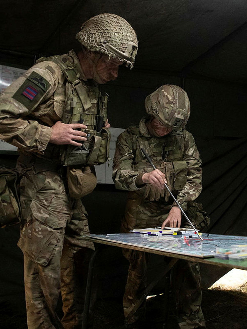 Three soldiers in camouflage uniforms plan a strategy around a table with maps and markers inside a dimly lit tent.