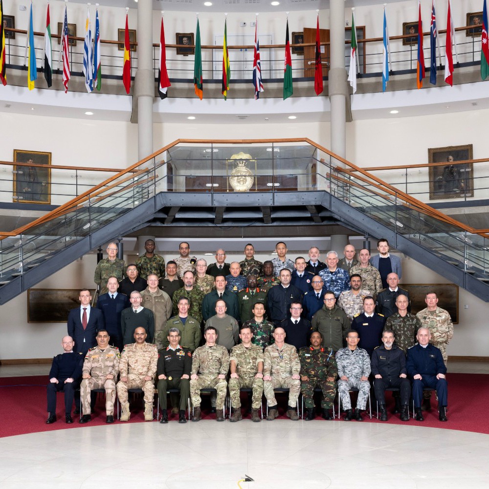The whole International Defence Attache party in a group event photo in the forum surrounded by international flags above them.