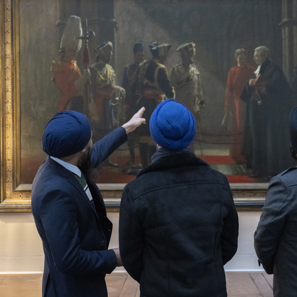 Three Sikh serving personnel with their backs to the camera, wearing blue turbans, pointing at a painting called Homage to the Fallen