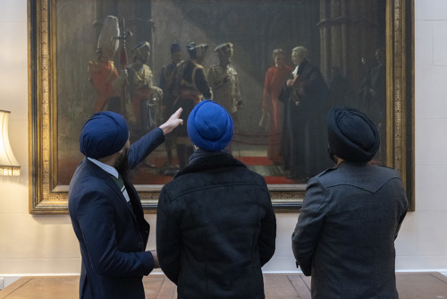 Three Sikh serving personnel with their backs to the camera, wearing blue turbans, pointing at a painting called Homage to the Fallen
