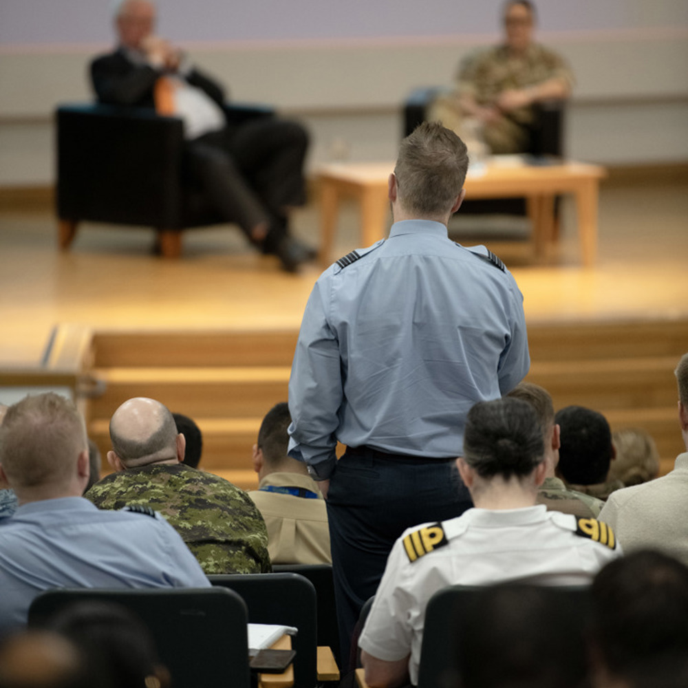 An RAF officer stood up among other audience members addressing a panel of speakers on stage. 