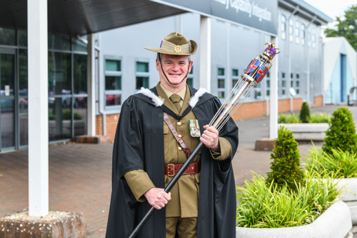 A man wearing a black robe and military uniform and carrying a mace posing in front of a building with green foliage to the right of him