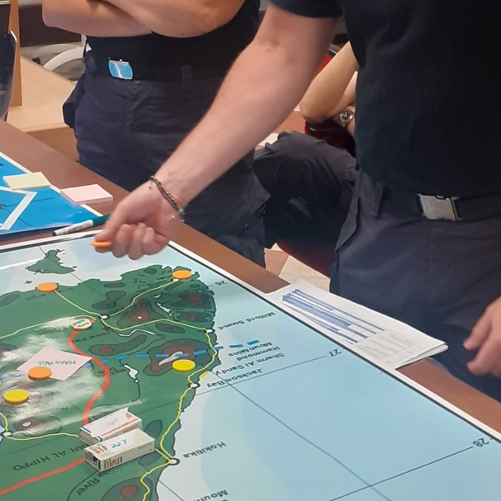 A group of people gathered around a table with a large map showing an island with marked locations. One person points at the map while others observe. A blue flag lies on the table, and a whiteboard and projector screen are visible in the background