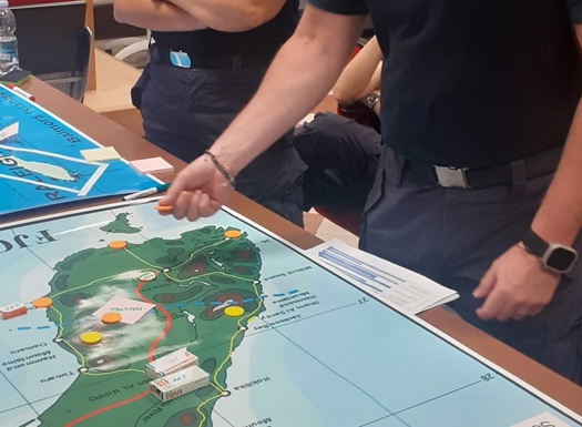 A group of people gathered around a table with a large map showing an island with marked locations. One person points at the map while others observe. A blue flag lies on the table, and a whiteboard and projector screen are visible in the background