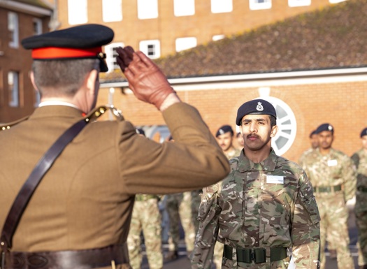 A military officer in a brown uniform salutes a soldier in camouflage during an outdoor ceremony.