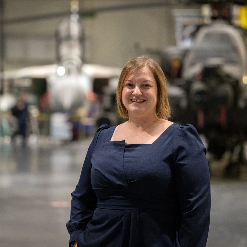 Smiling woman in a navy dress standing inside an aircraft hangar with military planes in the background.