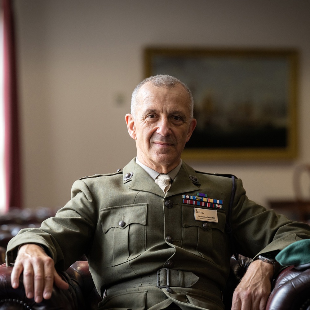 Senior military officer in formal uniform sitting confidently on a leather armchair in a classic room with framed paintings.
