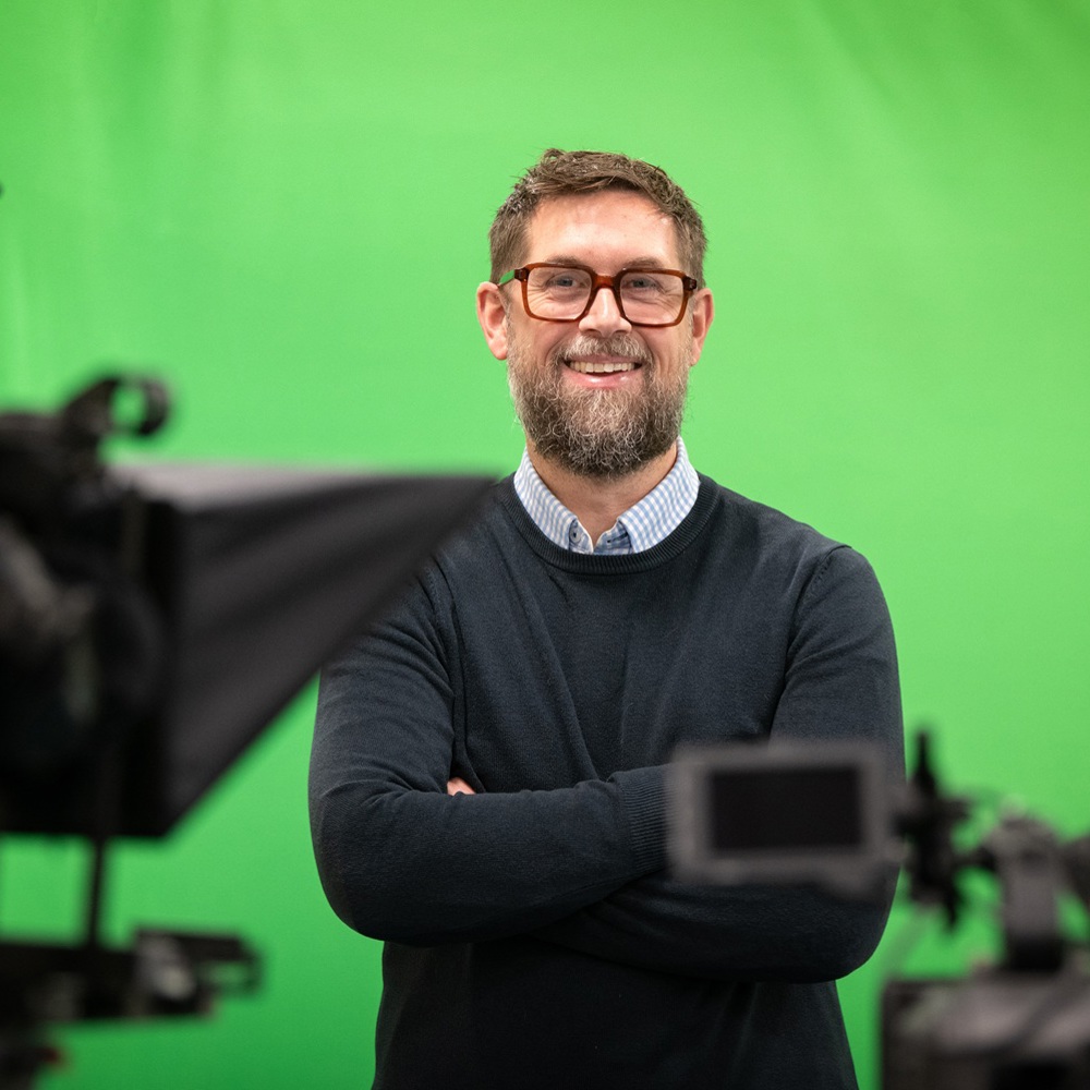 Man standing with arms crossed in front of a green screen, surrounded by professional video cameras and lighting equipment.