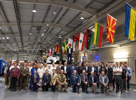 Large group of military and civilian personnel posing inside a hangar with a tank and international flags overhead.