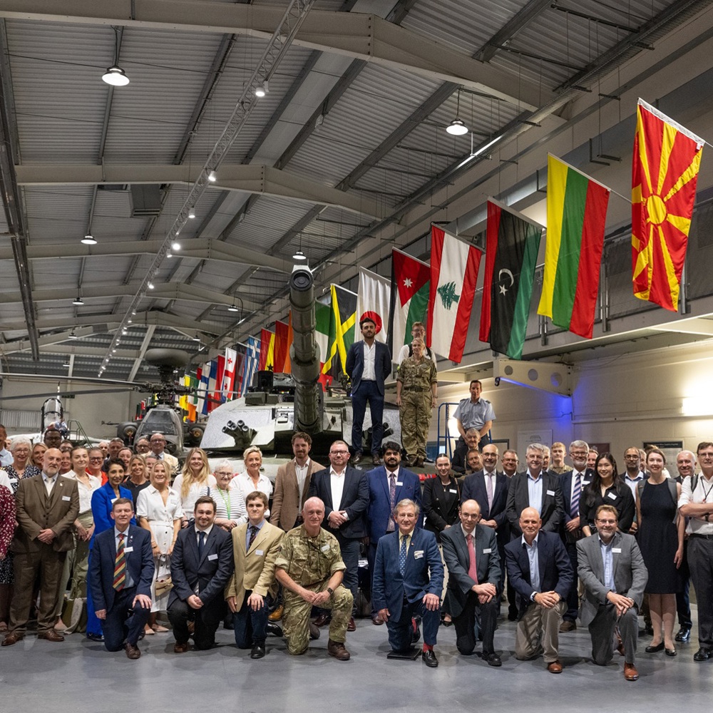Large group of military and civilian personnel posing inside a hangar with a tank and international flags overhead.
