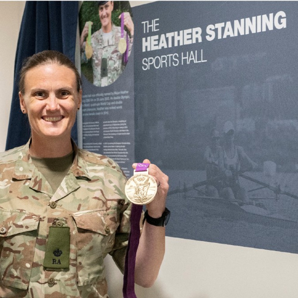 Heather Stanning holding her Olympic medals in front of her wall plaque inside the sports hall.