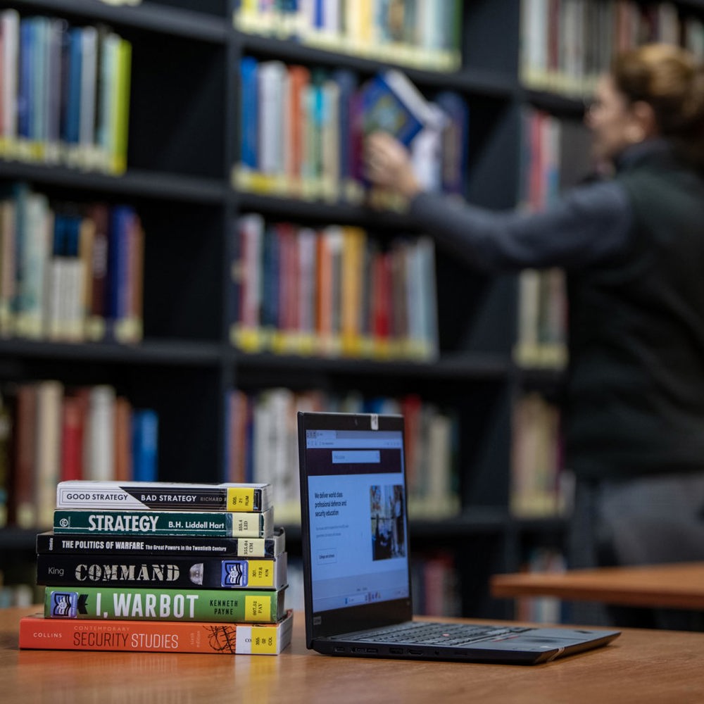 Stack of strategy and security books beside an open laptop on a library table with a person browsing shelves in the background.