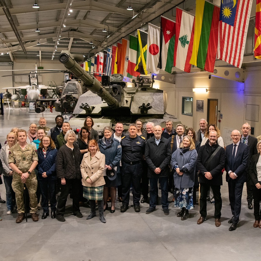 Group of diverse adults and military personnel posing in front of a large tank inside a museum hall with international flags overhead