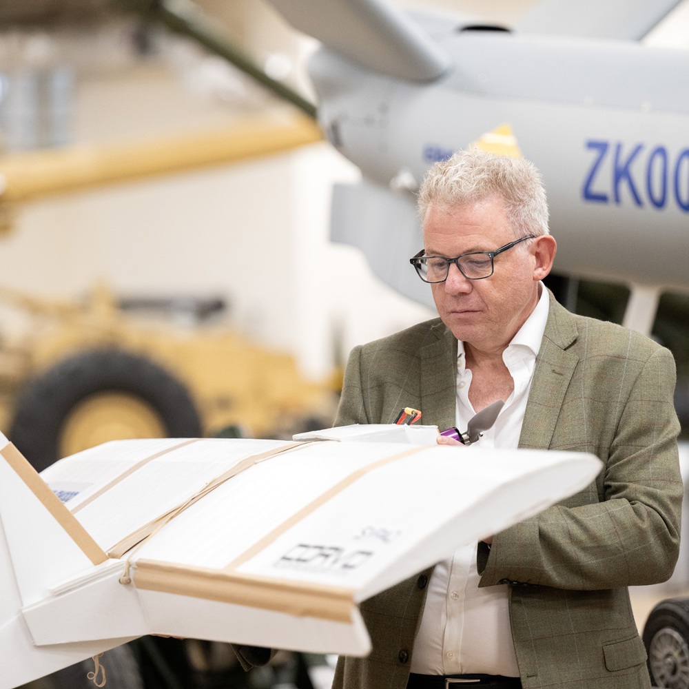 A person holds a small white drone labeled in front of a larger aircraft marked