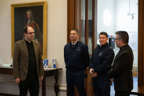 A Padre speaking to US Air Force officers inside a building with an oil painting in the background.