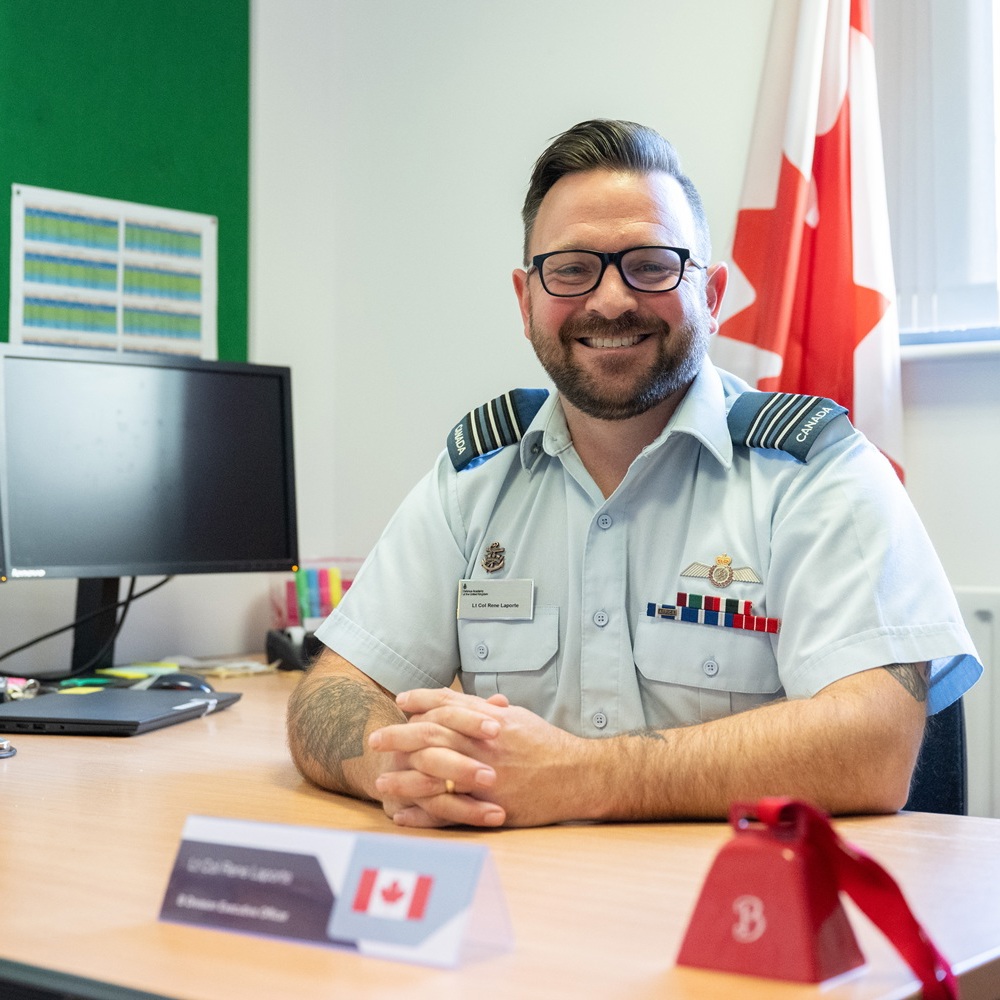 Canadian military officer in uniform sitting at a desk with folded hands and a Canadian flag behind him.