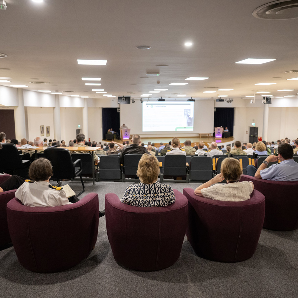 Audience seated in a large lecture theatre watching a presentation on a projector screen at the front.