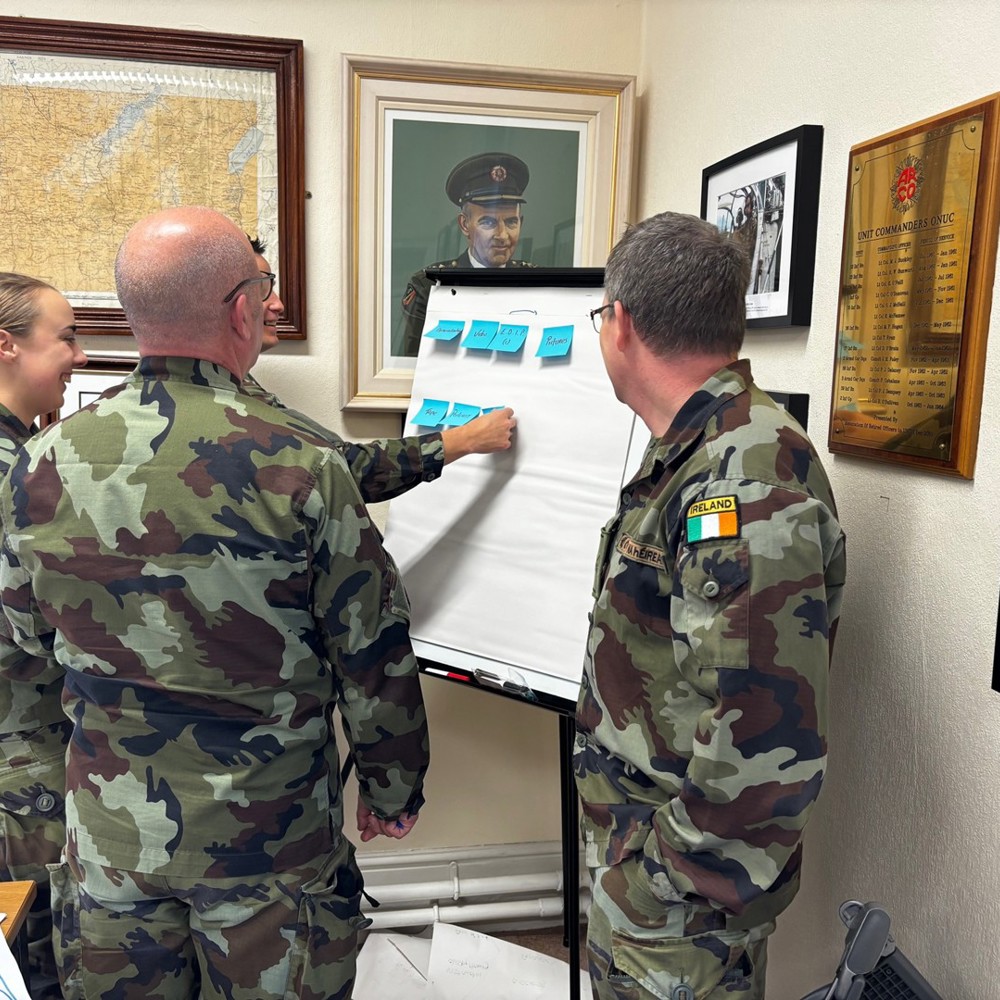 Four military personnel in camouflage placing sticky notes onto a flip chart. They are standing in front of various paintings and plaques