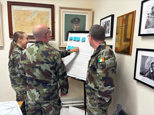 Four military personnel in camouflage placing sticky notes onto a flipchart. They are standing in front of various paintings and plaques