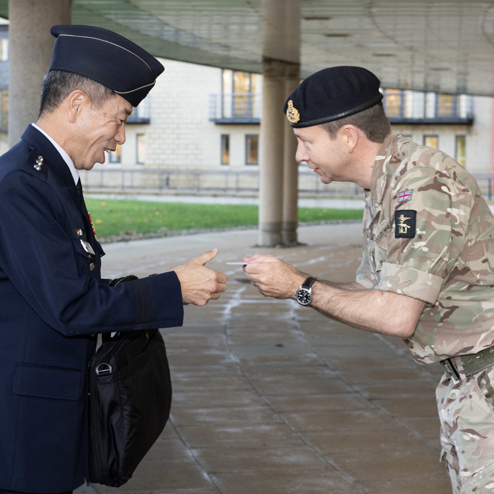 Major General Rowell, dressed in military working dress bows respectfully to his Japanese counterpart, whilst presenting him with a business card, as both men smile.