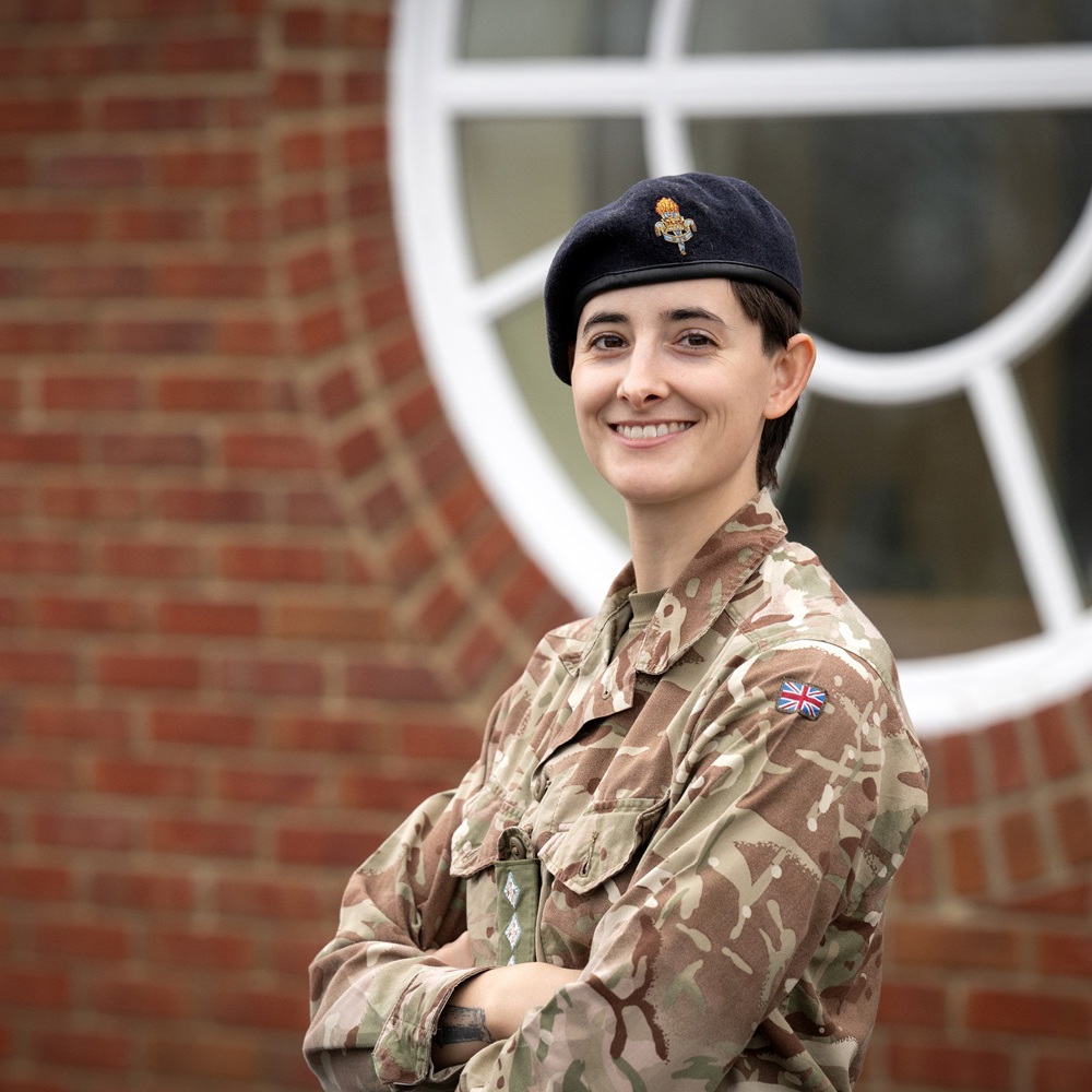 Soldier in camouflage uniform and black beret standing with arms crossed in front of a brick wall and circular window.