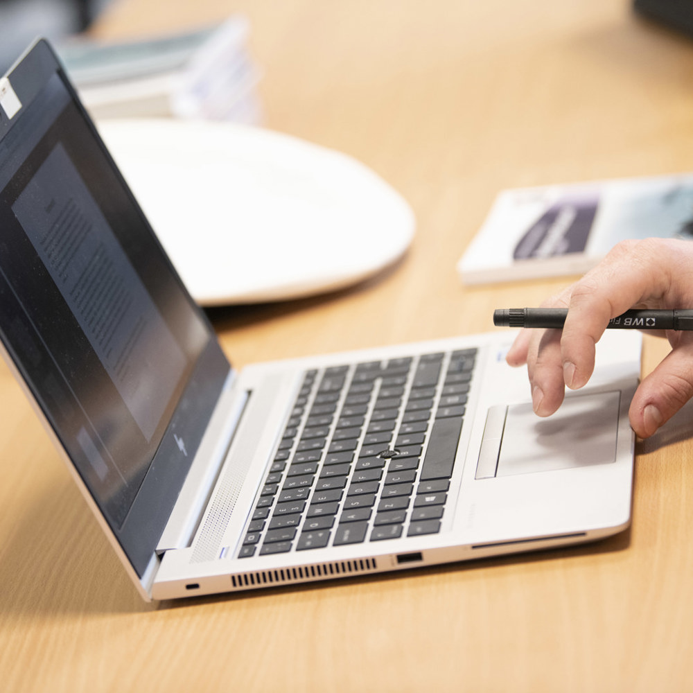 A laptop on a desk with a person using one hand to use the tracker pad. In their hand they hold a pencil