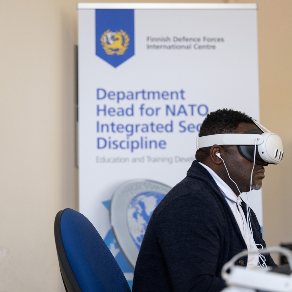 Person wearing a virtual reality headset and earphones seated in front of a Finnish Defence Forces International Centre banner.