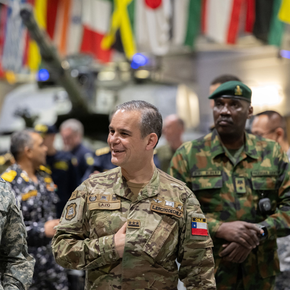 Military officers in different national uniforms walking in a large hall and smiling with international flags hung in the background. 
