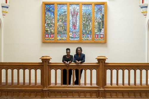 Two female members of the Gospel Choir stood looking over a balcony with a coloured glass pane window behind them.