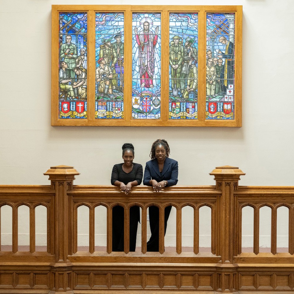 Two female members of the Gospel Choir stood looking over a balcony with a coloured glass pane window behind them.