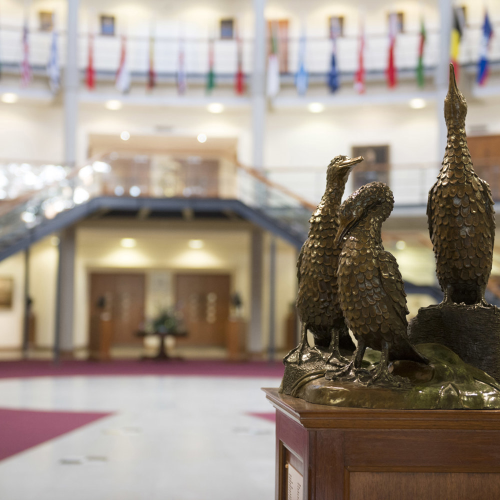 A gold statue of 3 cormorant birds inside a forum filled with multiple handing flags from around the world.