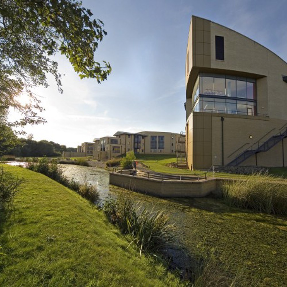 A ship-shaped wing of a building with a lake in the foreground and the sun setting behind. 