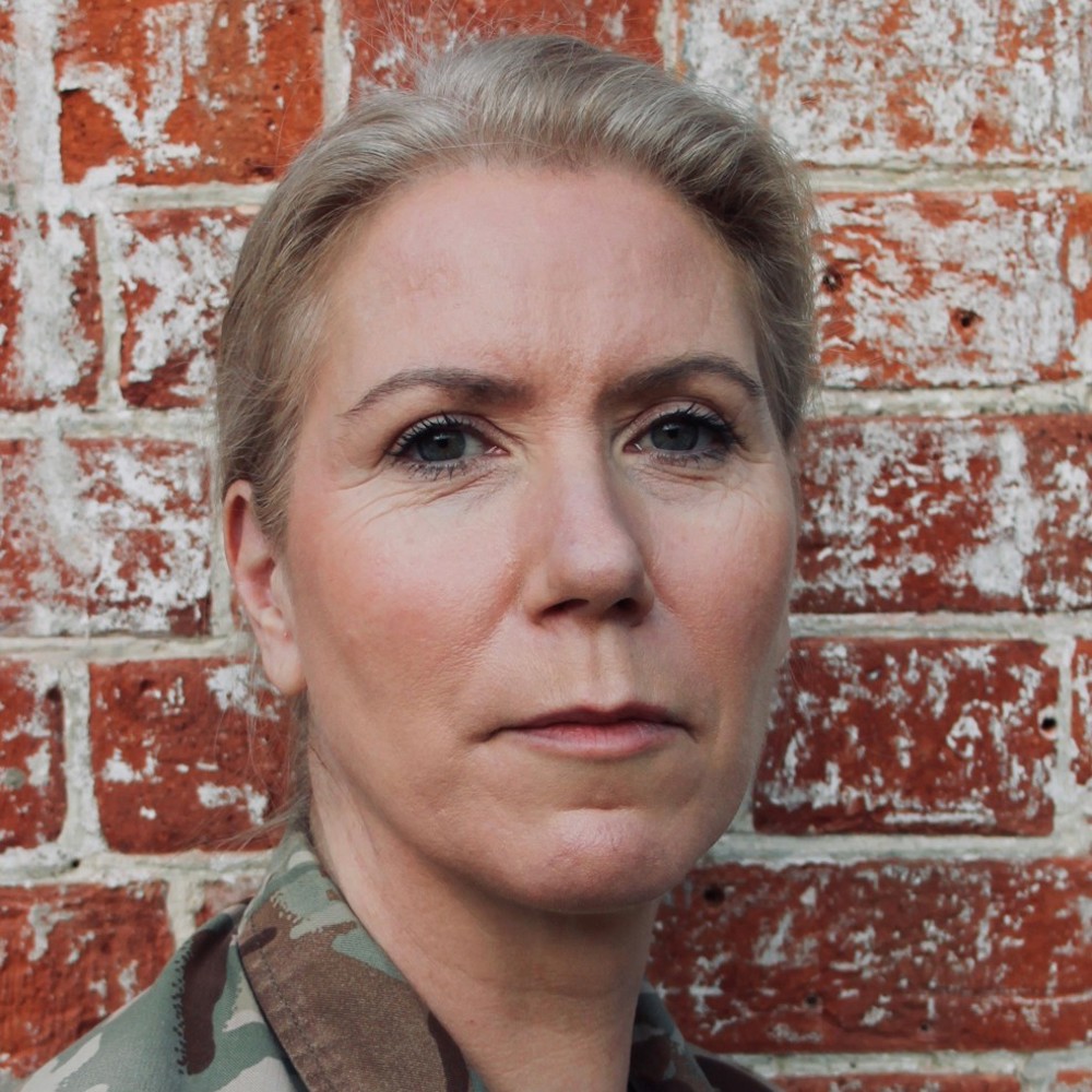 Person with blond hair in a bun wearing MTP uniform standing against a weathered red brick wall.