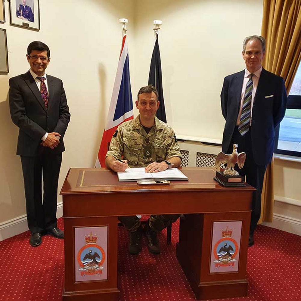 Army officer sat at a desk signing KIASS contract bookended by two individuals in suit and tie. 