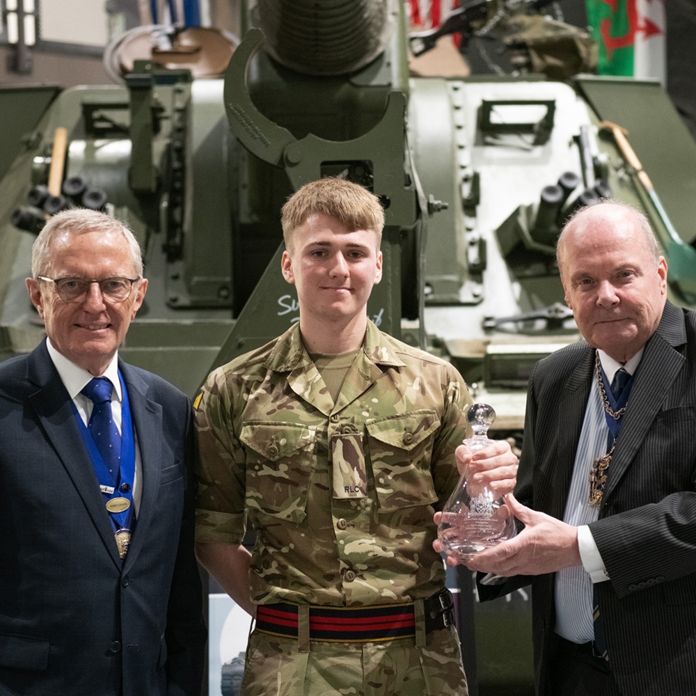 Three individuals stand in front of a military tank. The central figure wears a camouflage military uniform, while the two on either side are in formal attire with medals. The person on the right holds a glass trophy