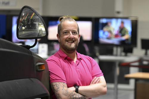 Civilian in pink shirt lent against an armoured vehicle with digital screens in the background.