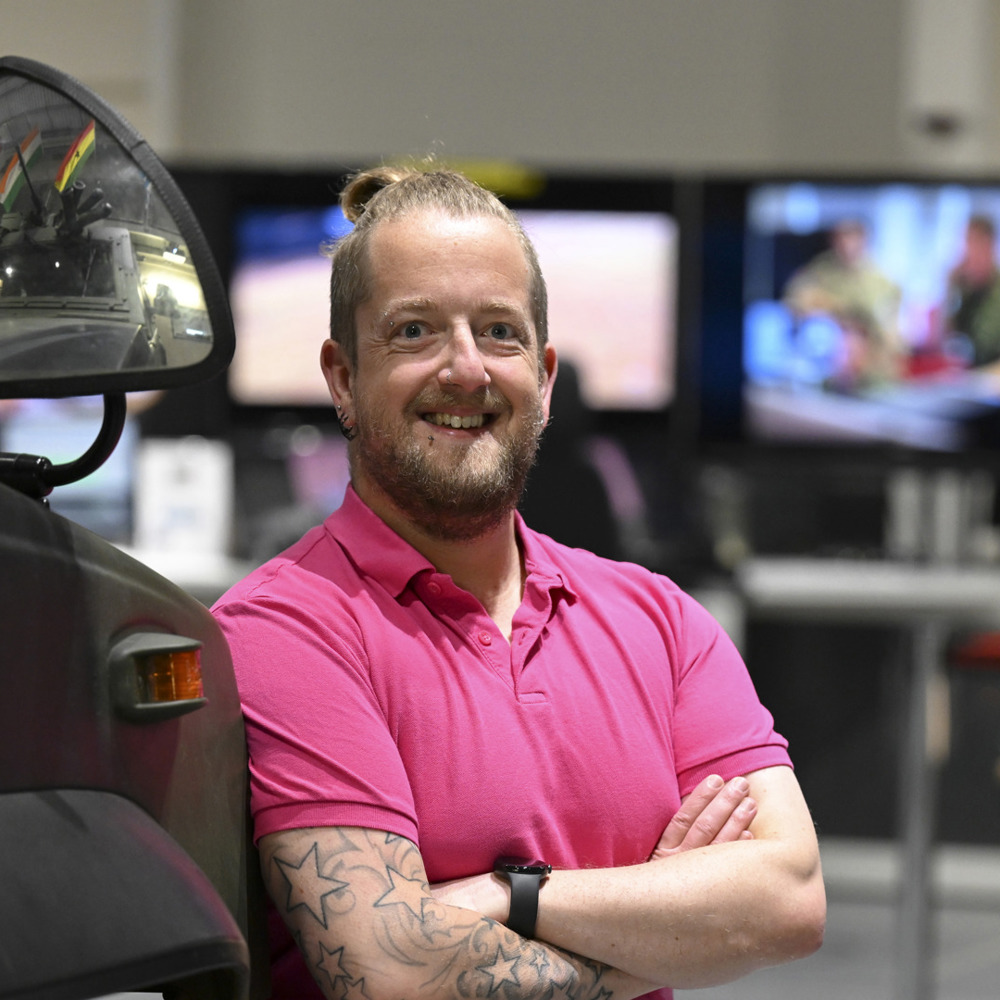 Civilian in pink shirt lent against an armoured vehicle with digital screens in the background.