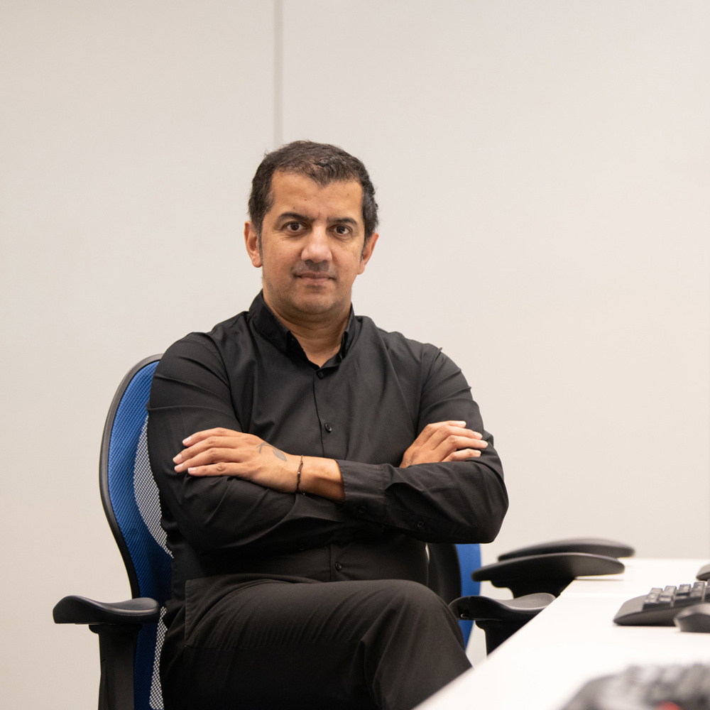 Man in black shirt sitting with arms crossed at a desk with multiple computer monitors in a minimalist office.