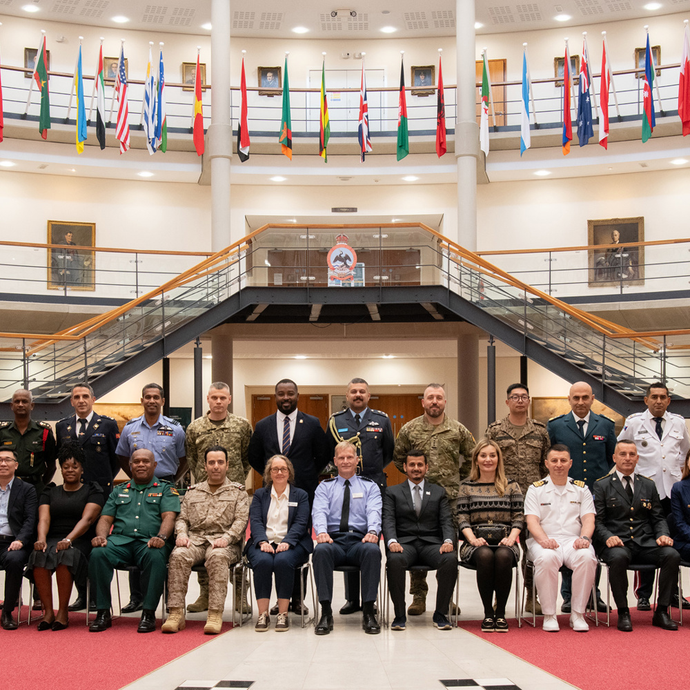 Group portrait of diverse military and civilian leaders seated and standing in a large hall with international flags overhead.
