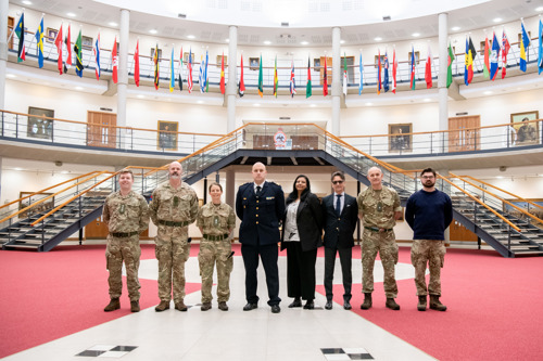 A group of British and Swedish military stood in a large hall with international flags hanging in the background.