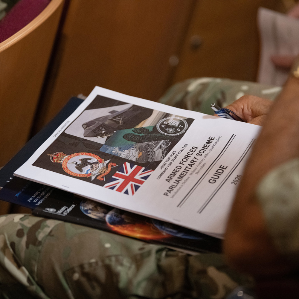 Close-up of a person in military uniform holding a guide titled 'British Forces Parliamentary Scheme' with various military insignias on the cover.