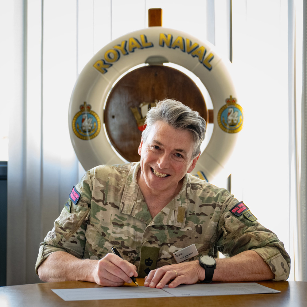 Military officer in camouflage uniform signing a document at a wooden table with a Royal Naval life ring displayed behind.