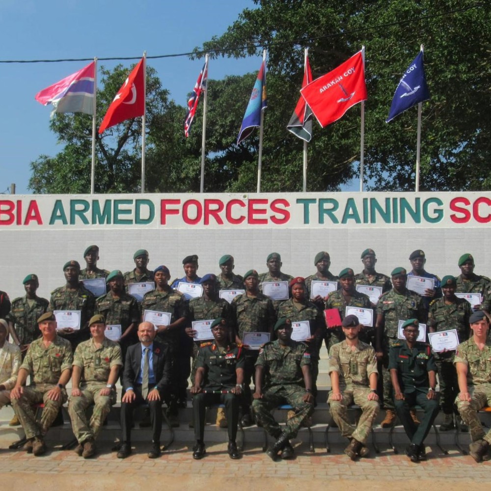 Group photo of Gambia Armed Forces trainees and instructors holding certificates in front of the training school sign and flags.