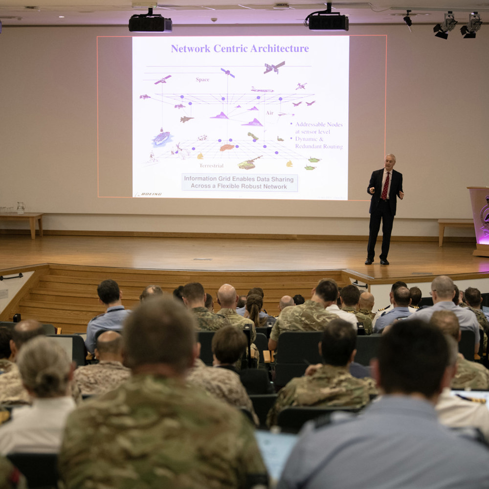 Civilian walking across a stage delivering a presentation to tri-service personnel.